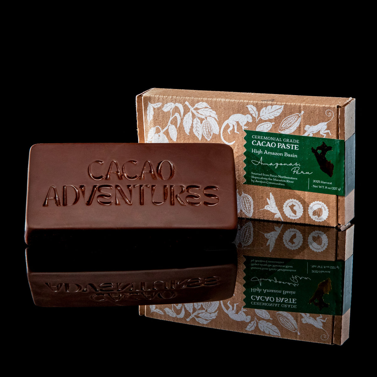A chocolate bar embossed with 'Cacao Adventures' sits in front of its kraft paper packaging box, which features white botanical illustrations and a green label, set against a solid black background with a reflective surface below.