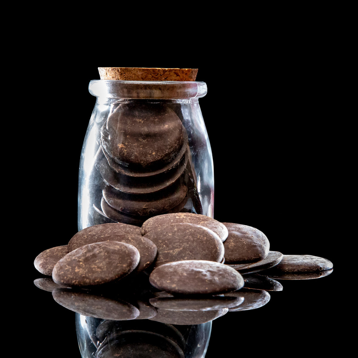 Dark brown smooth river stones arranged in and around a clear glass bottle with a cork stopper, photographed on a glossy black reflective surface against a black background.