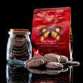 A red stand-up pouch of Encinitas California Cacao Adventures cocoa paste stands next to a small clear glass jar filled with round dark cacao discs, with several cacao discs scattered in front on a black reflective surface.