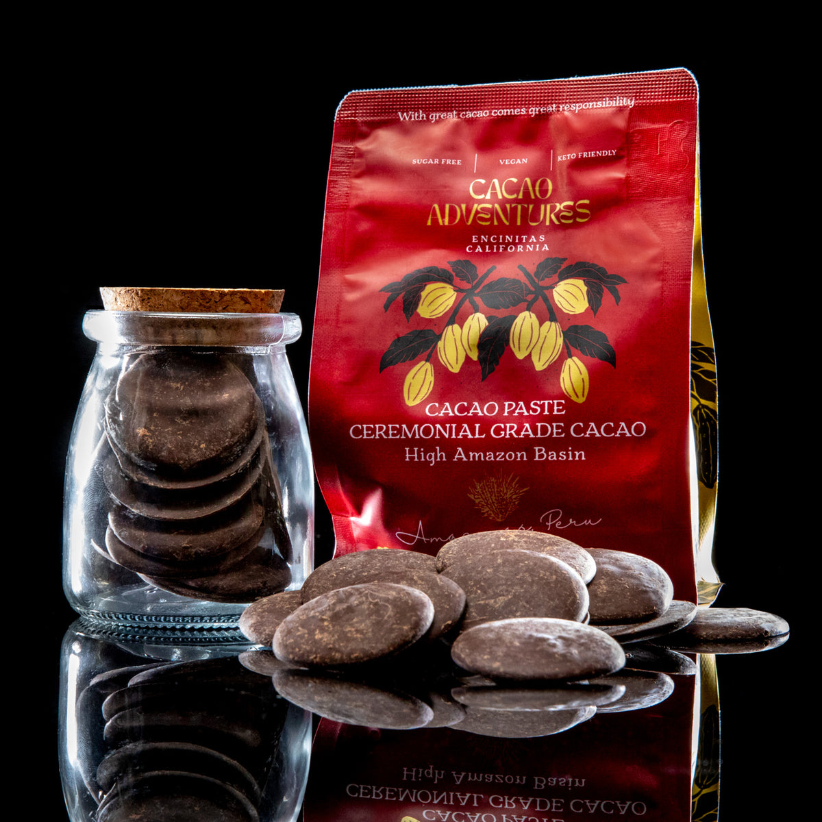 A red stand-up pouch of Encinitas California Cacao Adventures cocoa paste stands next to a small clear glass jar filled with round dark cacao discs, with several cacao discs scattered in front on a black reflective surface.