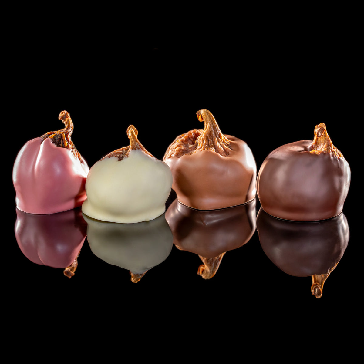 Four gourmet chocolate-covered confectionery items arranged in a row on a reflective black surface against a solid black background. The items include pink, white, and two shades of brown chocolate coating with caramelized tops.