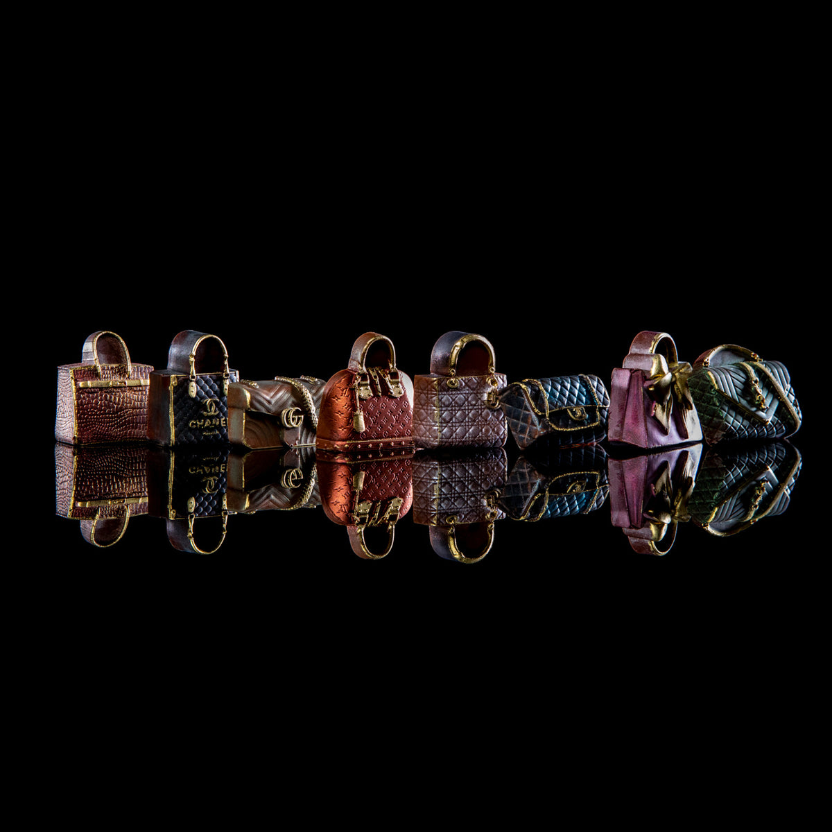 A row of miniature handbag-shaped jewelry charms or beads displayed on a glossy black reflective surface. The items include textured faux-leather and quilted fabric designs in pink, blue, brown, and green tones, each with gold-tone hardware.