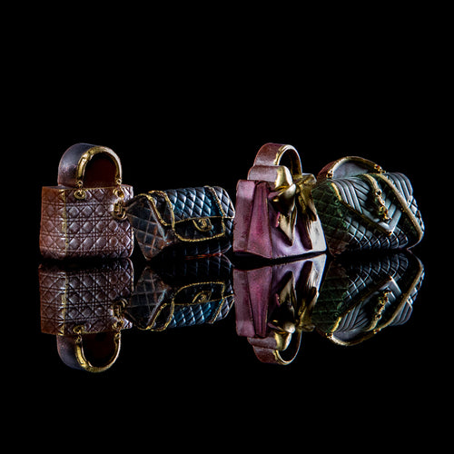 Three miniature quilted handbag-shaped objects in brown, blue, and pink, arranged on a glossy black reflective surface against a black background.