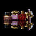 Four luxury perfume bottles arranged on a glossy black reflective surface against a black background. From left to right: a dark mauve Gucci Flower bottle with gold trim and cap, a lavender Versace bottle with a gold dome cap, a deep red CHANEL perfume bottle with gold detailing, and a lavender Versace bottle tied with a gold ribbon.