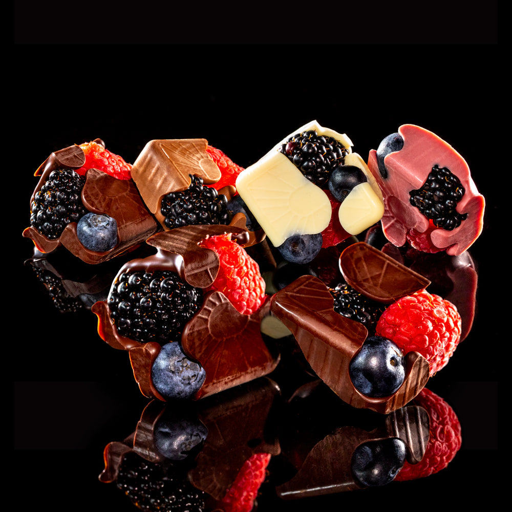 A heart-shaped arrangement of gourmet chocolate pieces decorated with fresh berries, including blackberries, raspberries, blueberries, and a white chocolate piece, set against a solid black background.