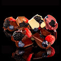 A heart-shaped arrangement of gourmet chocolate pieces decorated with fresh berries, including blackberries, raspberries, blueberries, and a white chocolate piece, set against a solid black background.