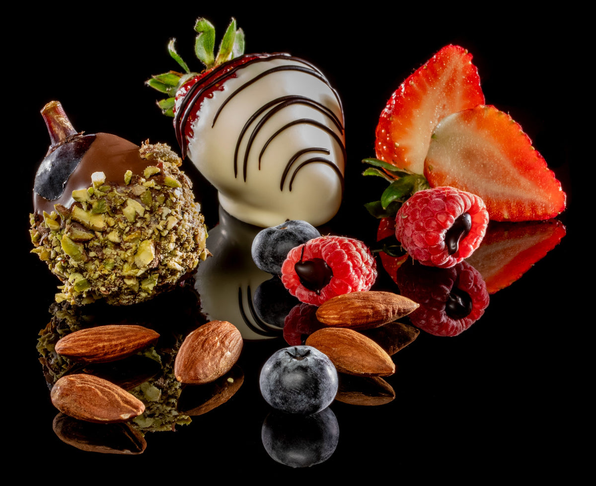 A styled arrangement of chocolate-covered berries and nuts on a glossy black reflective surface. The set includes a white chocolate strawberry with dark drizzle, a pistachio-dusted chocolate acorn, sliced strawberries, raspberries, blueberries, and almonds, all against a solid black background.