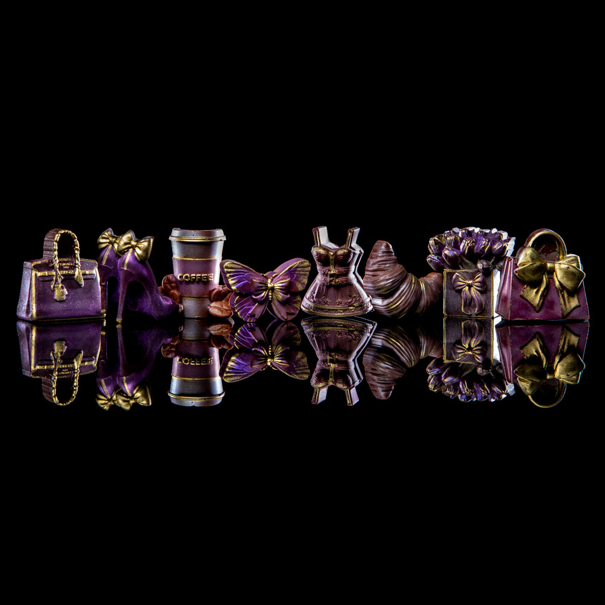 A set of miniature fashion accessories figurines in purple and gold, including handbags, a high-heel shoe, a coffee cup, a dress, and a flower, arranged on a reflective black surface against a black background.