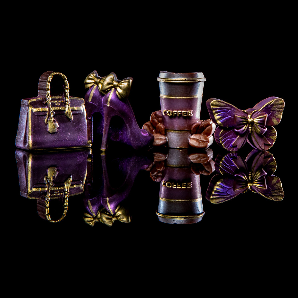 A set of miniature fashion and coffee-themed charms on a reflective black surface, including a purple handbag, high heel, coffee cup, butterfly, and flowers, all decorated with gold-tone detailing.