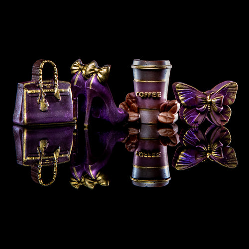 A set of miniature fashion and coffee-themed charms on a reflective black surface, including a purple handbag, high heel, coffee cup, butterfly, and flowers, all decorated with gold-tone detailing.