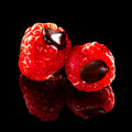 Two glossy red raspberries with dark chocolate centers, photographed on a black reflective surface.